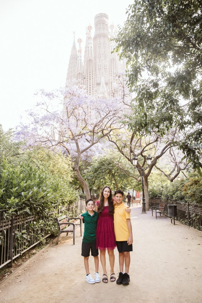 A woman stands between two children on a park path lined with trees, with the Sagrada Família basilica visible in the background. The group smiles at the camera, and the area is lush with greenery and benches.