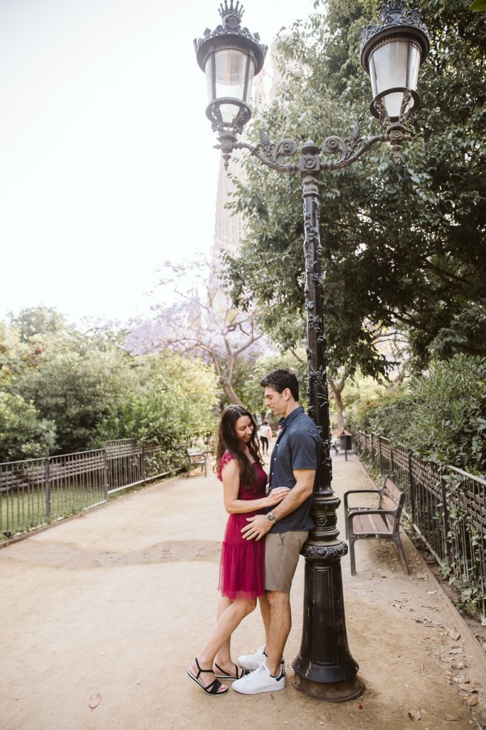 A couple stands close together under a tall black lamppost on a tree-lined park path. The woman wears a pink dress and sandals; the man wears a blue shirt, shorts, and white sneakers. Benches and greenery are in the background.