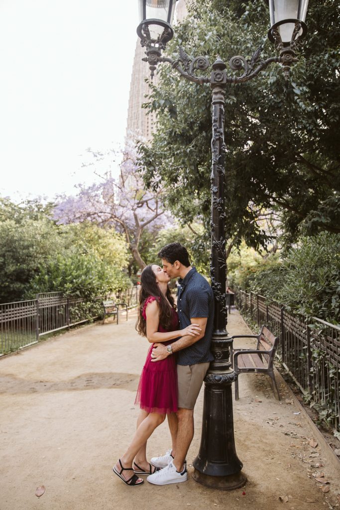A couple kisses under a vintage lamp post on a park path, surrounded by greenery and benches, with the man wearing a navy shirt and shorts and the woman in a pink dress and sandals.