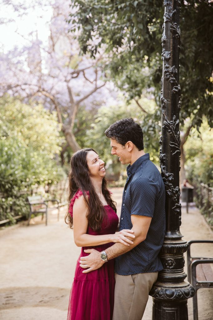 A couple stands close together in a sunlit park, smiling at each other. The woman in a red dress rests her hands on the man’s chest as he leans against a decorative lamppost surrounded by greenery and trees.