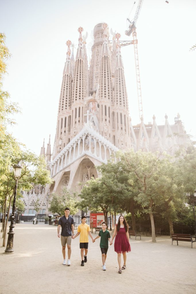 A family of four walks hand in hand in front of the Sagrada Família basilica in Barcelona, Spain, on a sunny day. The iconic church and a construction crane are visible in the background.