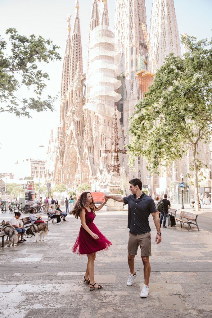 A couple dances joyfully on a sunny street in front of the Sagrada Família basilica in Barcelona, surrounded by trees, benches, and people walking or sitting nearby.
