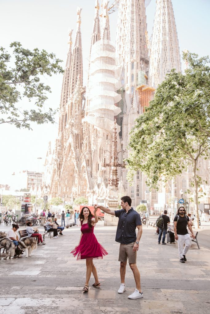 A couple dances joyfully in front of the Sagrada Familia in Barcelona on a sunny day, surrounded by people, trees, and dogs in the busy plaza.