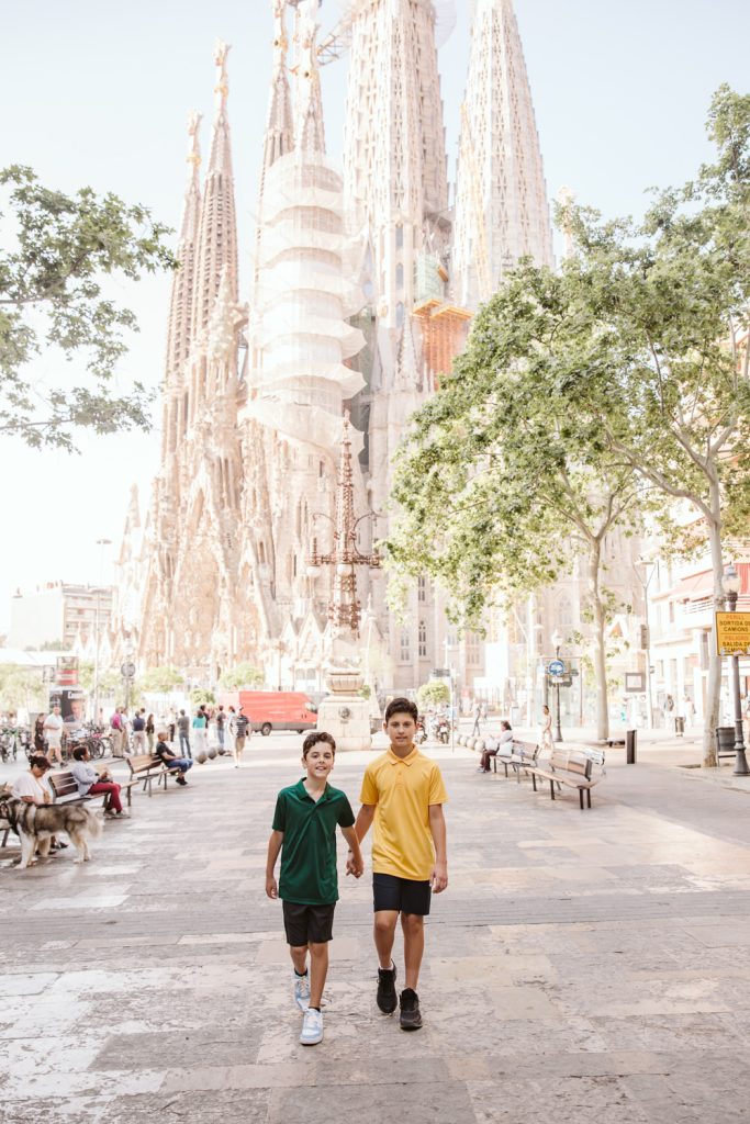 Two boys holding hands walk on a sunny street with the Sagrada Familia basilica in Barcelona visible behind them. Trees line the street, and people sit on benches along the sidewalk.