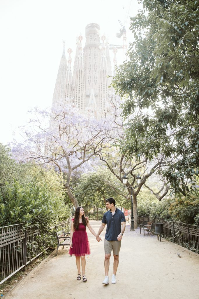 A couple holding hands walks down a tree-lined path, with the Sagrada Família basilica visible in the background. The woman wears a pink dress, and the man wears a blue shirt and shorts. The scene is bright and peaceful.