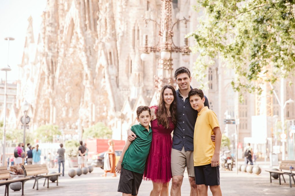 A smiling family of four poses together outdoors near the Sagrada Família in Barcelona. The parents stand in the center, with two boys in green and yellow shirts standing on each side. Trees and benches are visible nearby.