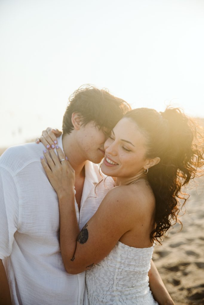 A couple embraces on a sandy beach at sunset, smiling and holding each other closely. The person on the left wears a white shirt, while the person on the right wears a white dress and has curly hair.