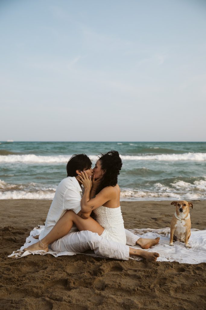 A couple in white clothing sits on a blanket kissing by the ocean, with waves in the background and a small tan dog sitting nearby on the sandy beach.