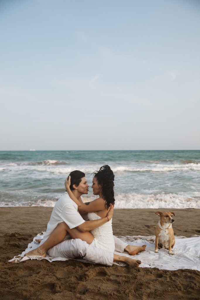 A couple in white clothing sits embracing on a blanket at the beach, facing each other and smiling. A brown dog sits nearby on the sand. The ocean and sky are visible in the background.