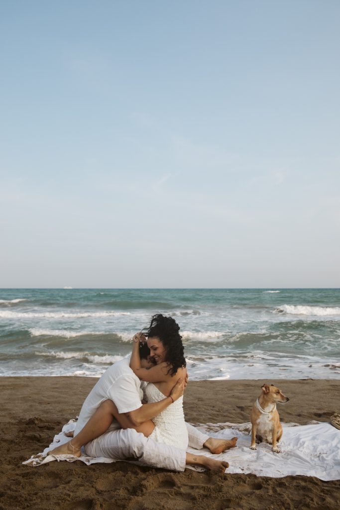 Two people embrace lovingly while sitting on a blanket at the beach, with waves in the background and a dog sitting beside them on the sand under a clear sky.