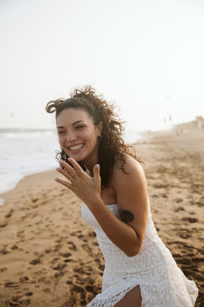 A woman in a white dress smiles and shows her hand to the camera while standing on a sandy beach near the ocean, with gentle waves and a hazy sky in the background.