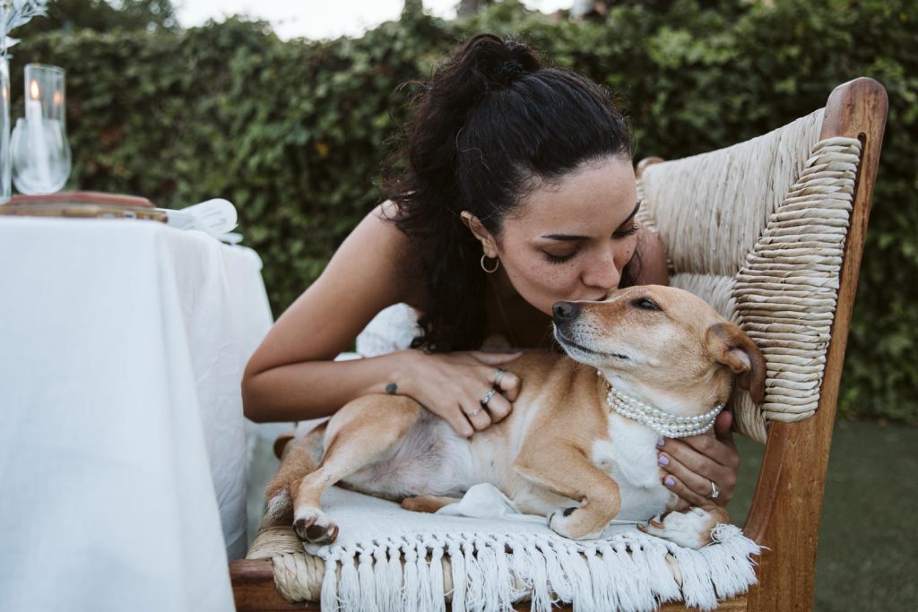 A woman kisses a small dog wearing a pearl necklace as it lies on a woven chair next to a table outdoors, with greenery in the background.
