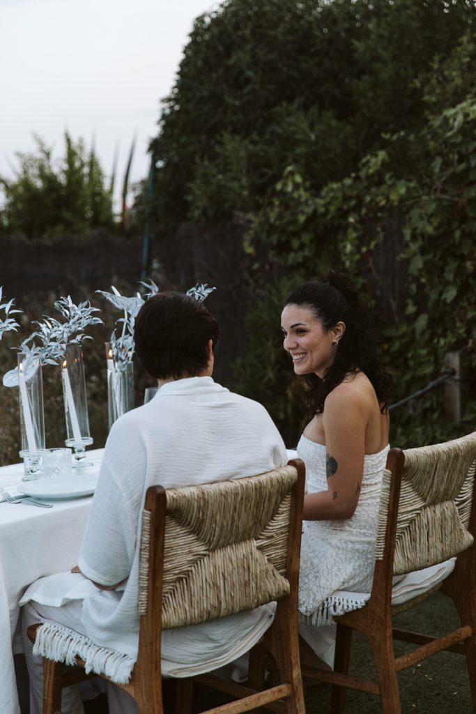 Two people sit side by side at an outdoor table decorated with glass vases and silver foliage. One person, facing away, wears white. The other, smiling, wears a white dress. Greenery surrounds the scene.