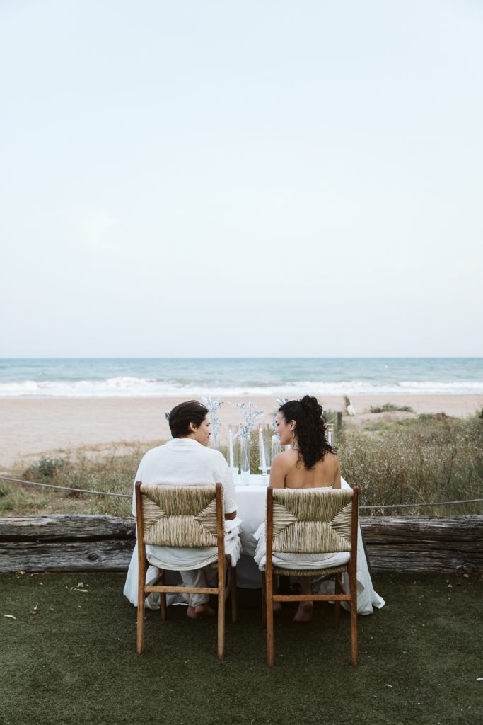 A couple sits at a decorated table on a grassy area by the beach, facing the ocean. They are dressed in white and surrounded by natural scenery, with waves and sky in the background.