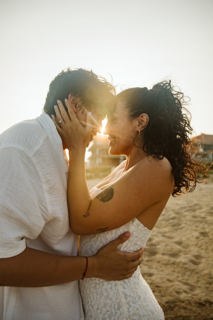 A couple embraces on a sandy beach at sunset, smiling and touching foreheads closely. The woman holds the mans face with both hands as sunlight shines between them, creating a warm, intimate atmosphere.