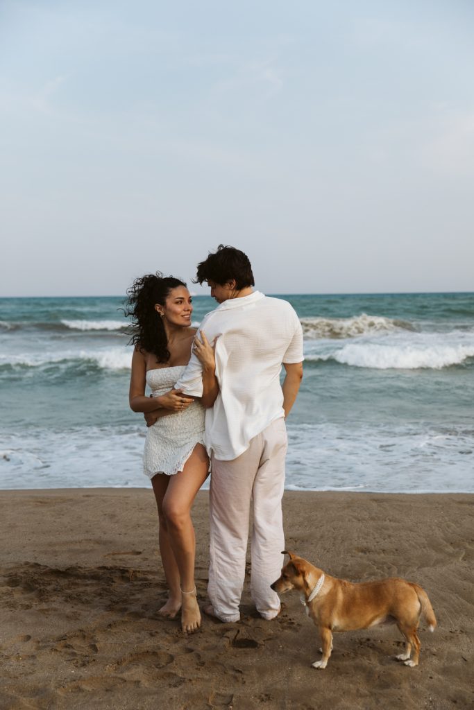 A couple stands on a sandy beach facing each other, with the ocean waves behind them. The woman wears a white dress, the man wears a white shirt and light pants, and a small brown dog stands by their feet.