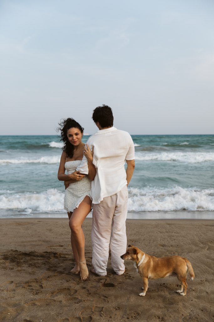 A couple stands barefoot on a sandy beach near the ocean, with the woman smiling at the camera and the man facing away. A small brown dog stands by their feet. Waves and a cloudy sky are in the background.