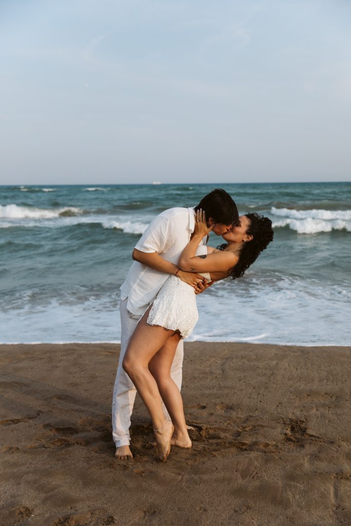 A couple dressed in white shares a passionate kiss and embrace on a sandy beach, with ocean waves in the background and clear sky above.