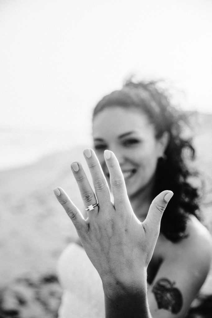 A woman on a beach smiles and holds her hand up toward the camera, showing off a ring on her finger. The image is in black and white and her face is softly focused in the background.