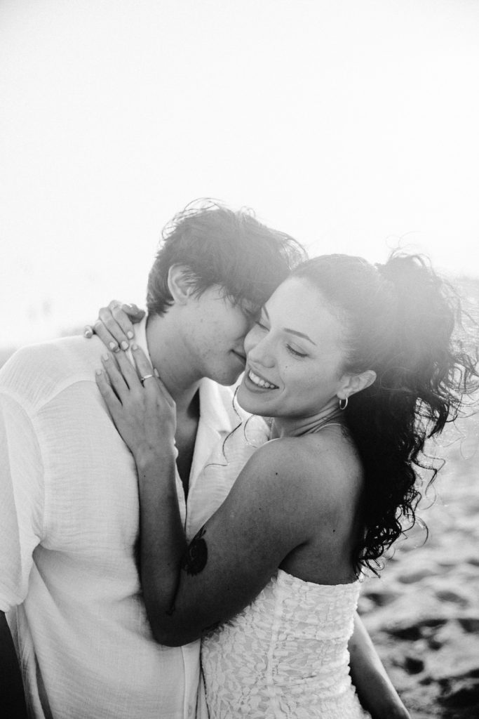 A couple embraces on a beach, smiling and enjoying a tender moment. The woman wears a strapless dress and has curly hair, while the man wears a light shirt. The photo is in black and white with a soft, bright background.