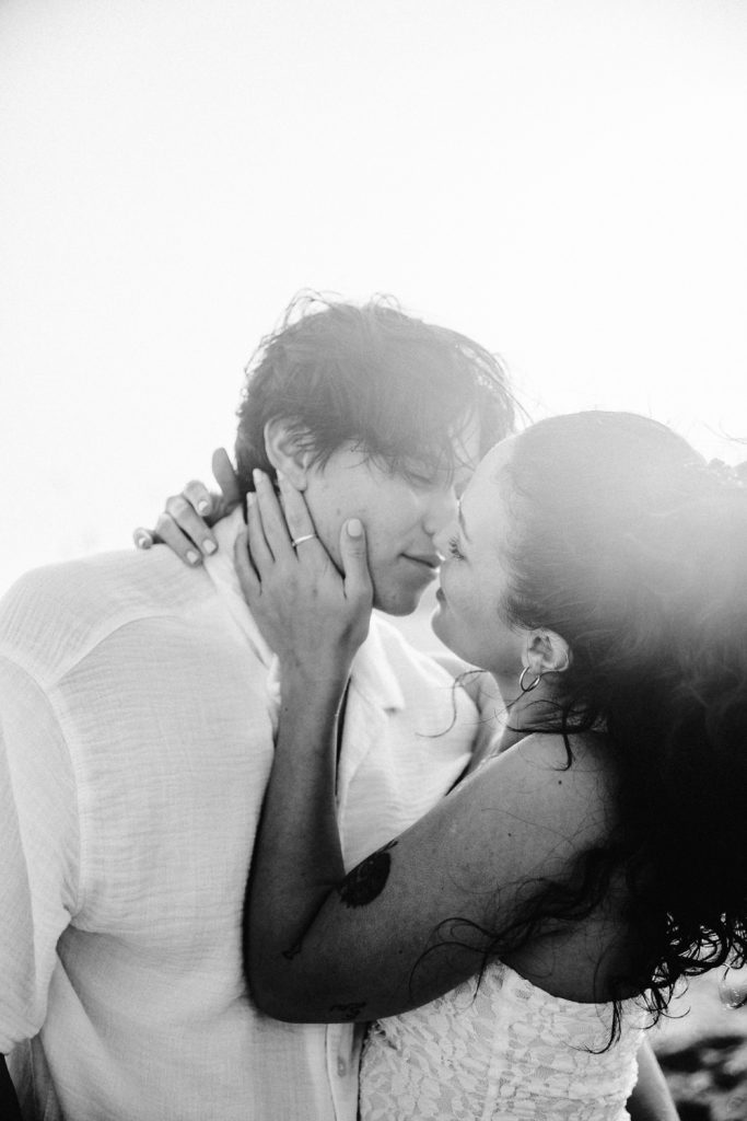A couple stands close, embracing and about to kiss. The woman gently holds the mans face with both hands. The image is black and white, with soft lighting creating a romantic and intimate atmosphere.