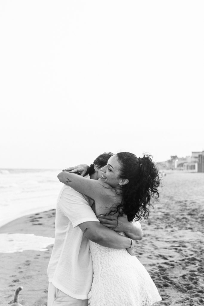 A joyful woman with curly hair hugs a person tightly on a sandy beach, both smiling. The ocean and beach houses are visible in the background. The photo is in black and white.