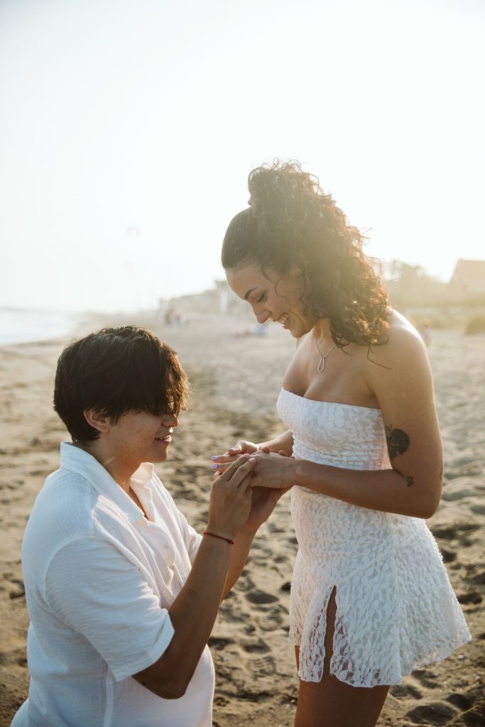 A person kneels on the beach, holding a ring as they propose to a smiling woman in a white dress. The sun is shining, and both appear happy and emotional.