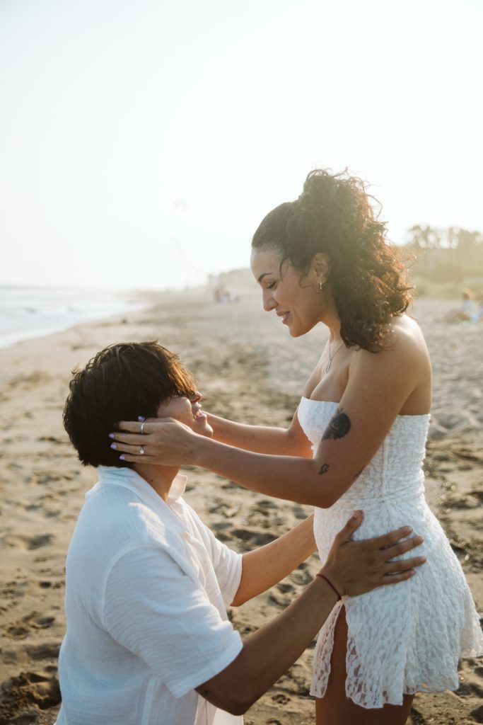 A couple on a sandy beach at sunset; the woman smiles and holds the man’s face while he kneels, gazing up at her. Both wear white, and the scene is warm and romantic with soft sunlight.