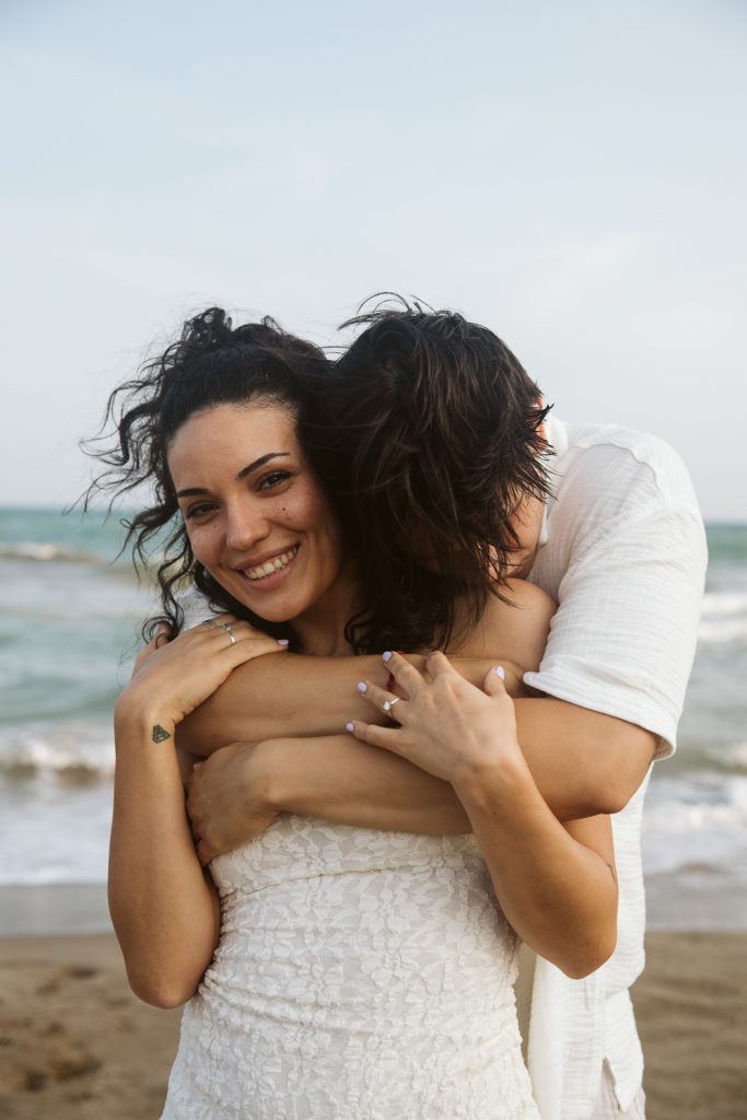 A smiling woman in a white dress stands on the beach while another person hugs her from behind, resting their head on her shoulder. The ocean and sky are visible in the background.